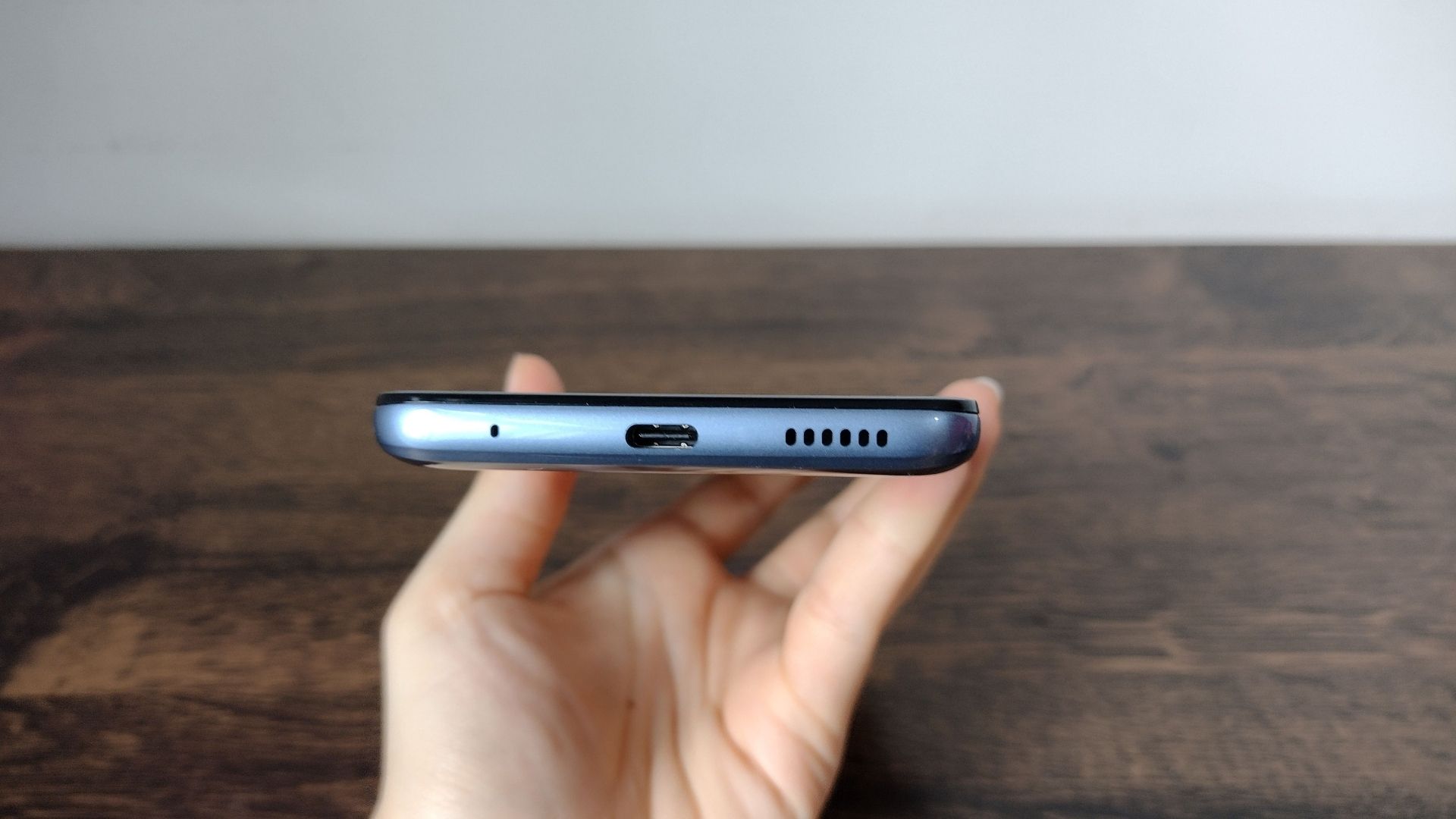 Imagem mostra mão segurando Moto G60 e exibindo parte de baixo do celular, onde fica a entrada para o carregador e uma mesa de madeira no fundo